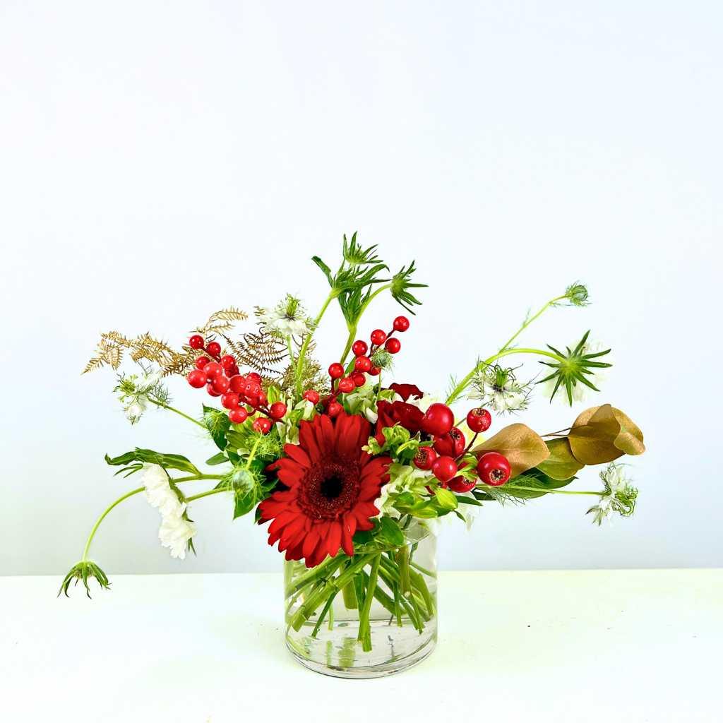 Christmas-themed Holly Jolly Blossom Flower Arrangement with red gerberas, faux berries and greenery in a small glass vase - Fabulous Flowers