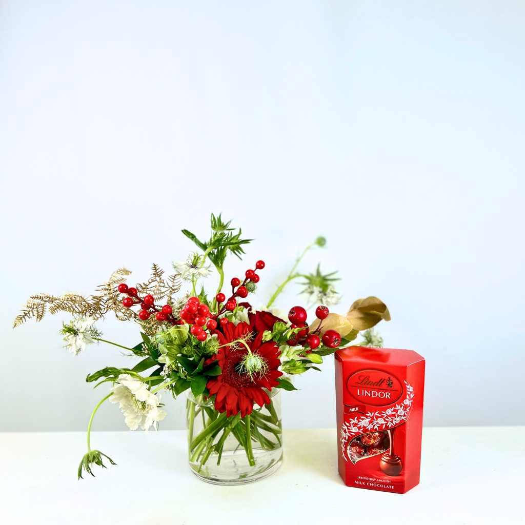 Christmas-themed Holly Jolly Blossom Flower Arrangement with red gerberas, faux berries and greenery in a small glass vase - Fabulous Flowers