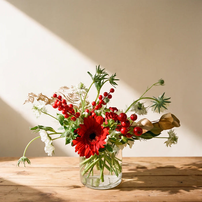 Small Christmas flower arrangement of red gerberas, red berries, white scabiosa and gold fern in a glass vase on a sunlit pine table, Fabulous Flowers South Africa