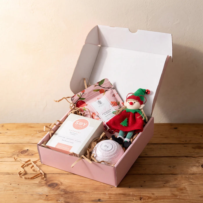 Open pale pink gift box with bath salts, a soy candle, a pink bath bomb and a felt elf ornament on a sunlit pine table, Fabulous Flowers South Africa