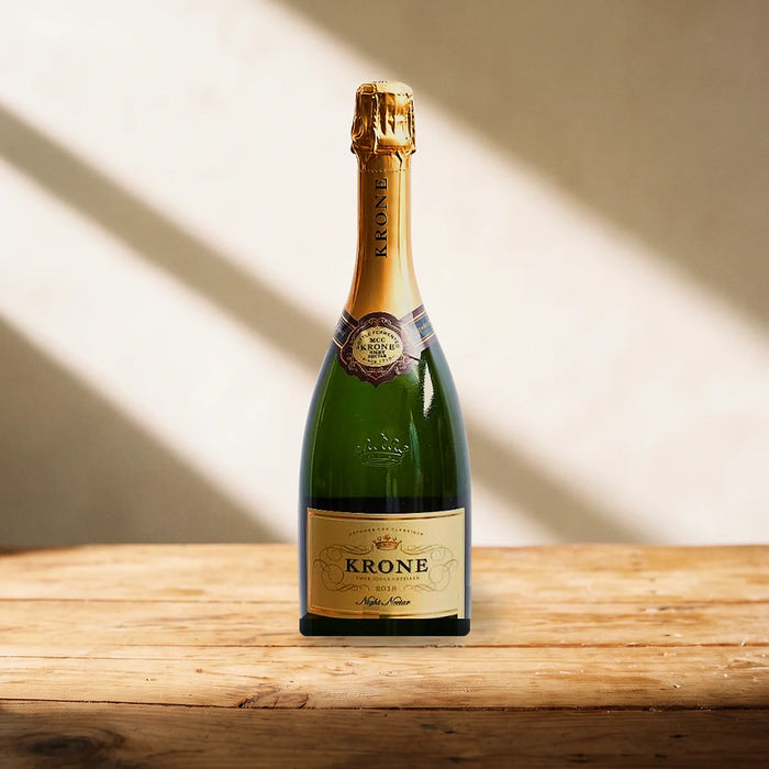 Bottle of Krone Night Nectar Cap Classique with a gold foil neck and gold crown label standing on a pine table in late-afternoon sunlight, Fabulous Flowers South Africa