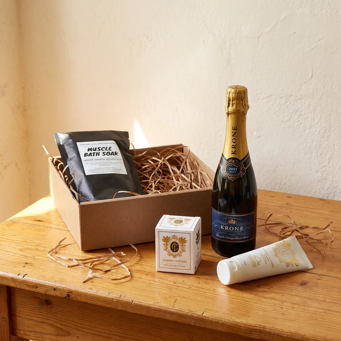 Kraft gift box with a black muscle bath soak pouch, beside a half bottle of Krone, a Cape Island candle box and hand cream on a pine table, Fabulous Flowers South Africa