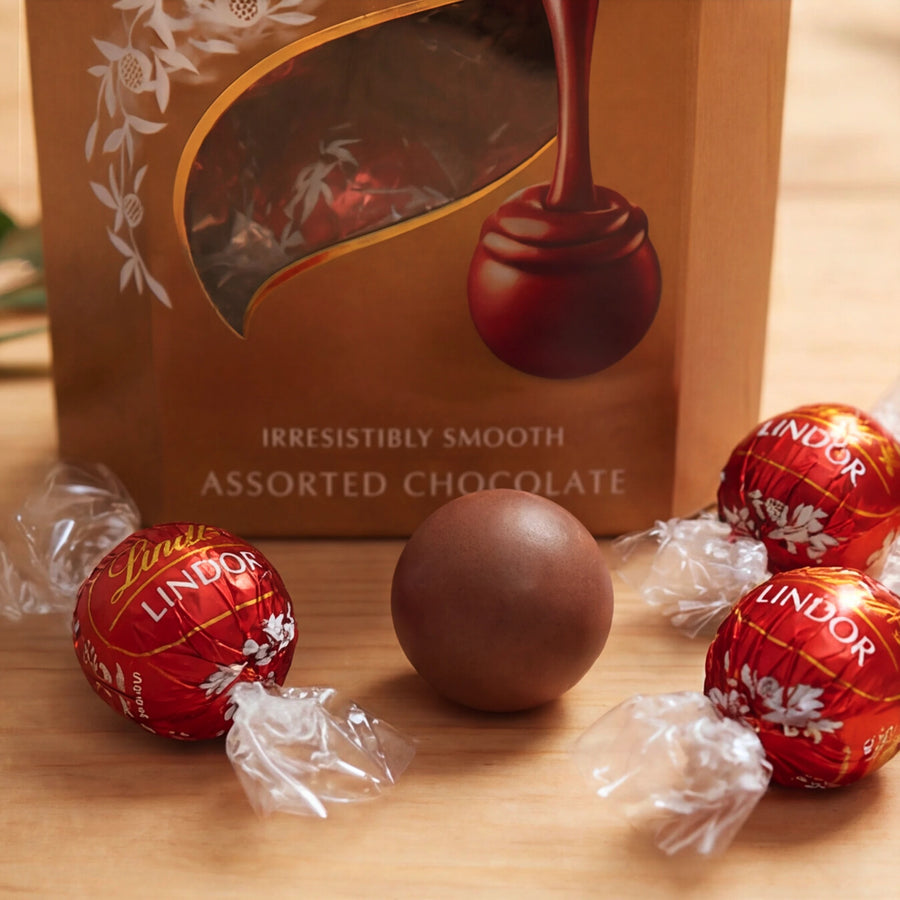 Close-up of Lindt Lindor Assorted Chocolate box with red foil truffles and an unwrapped smooth milk chocolate ball