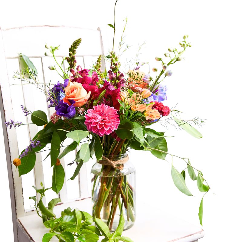 Jar flower arrangement of colorful flowers in a clear vase on a white chair.