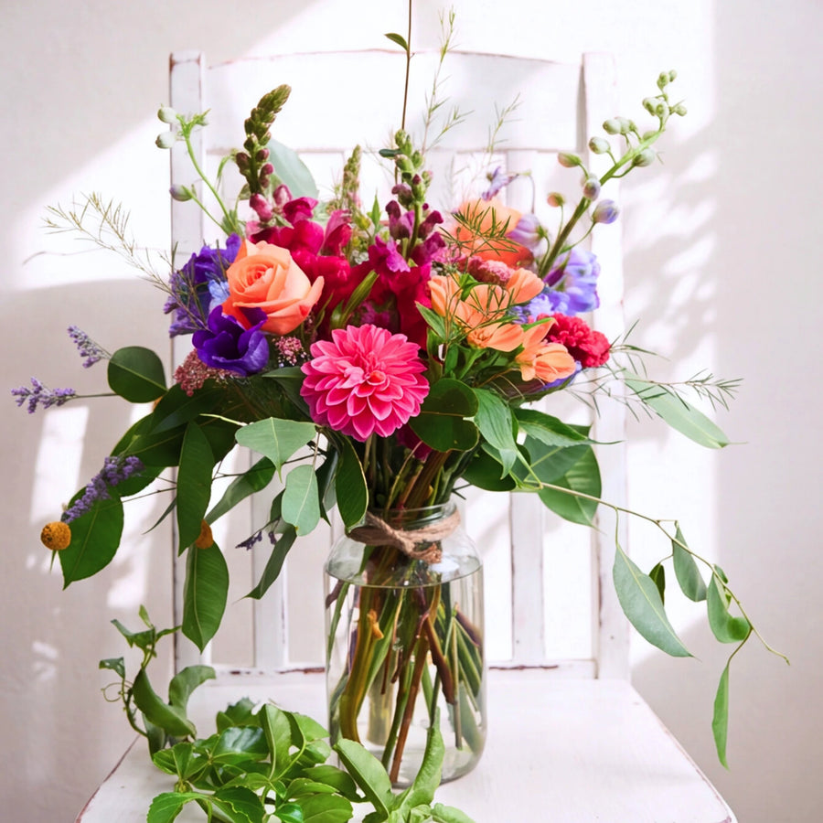 A vibrant, multicolored wildflower bouquet arranged in a clear glass jar with a twine bow, set against a white rustic wooden chair with soft sunlight and shadows.
