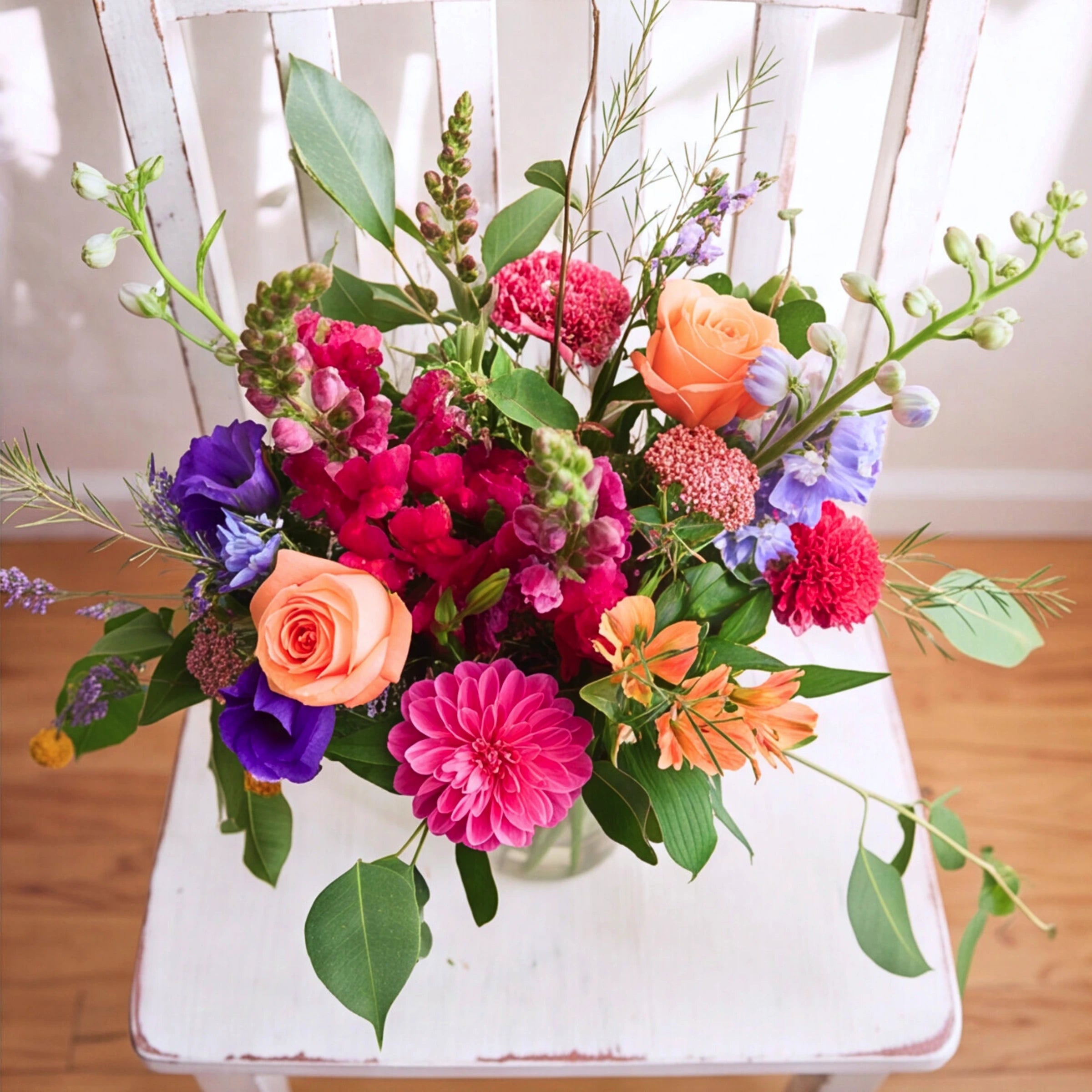A vibrant, multicolored floral arrangement featuring pink, orange, purple, and red flowers with green foliage, displayed on a distressed white wooden chair.