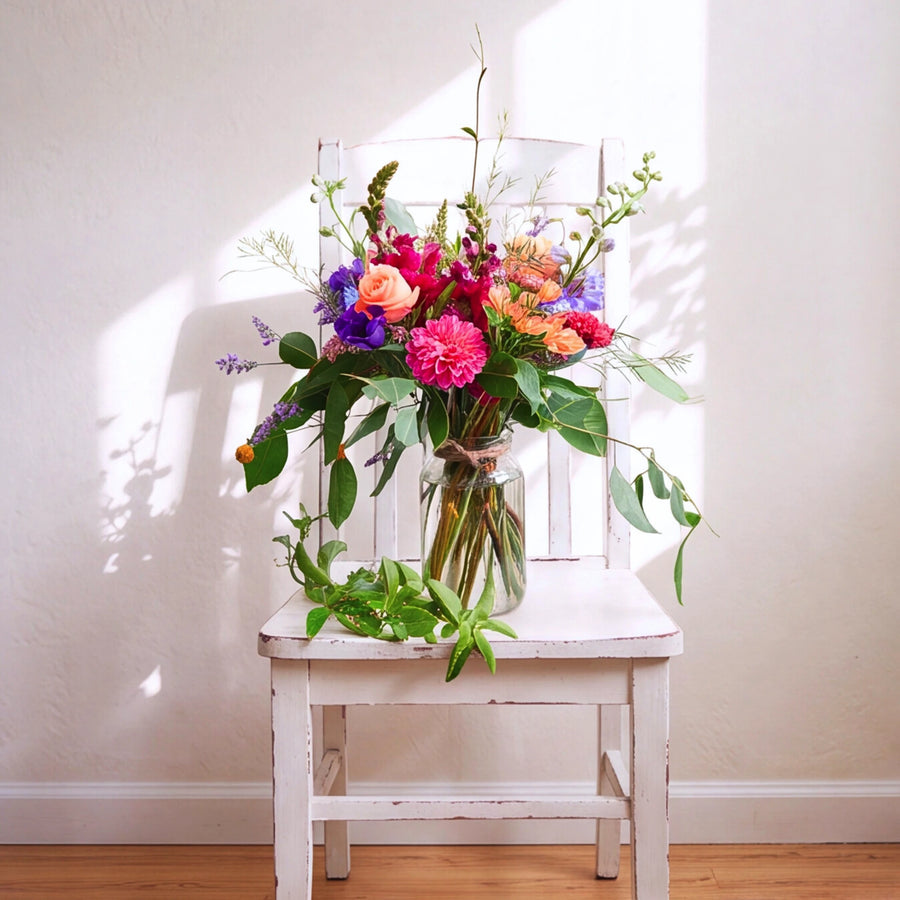 A vibrant, multicolored wildflower bouquet in a clear glass jar vase, styled on a distressed white wooden chair against a textured cream wall with soft window light shadows.