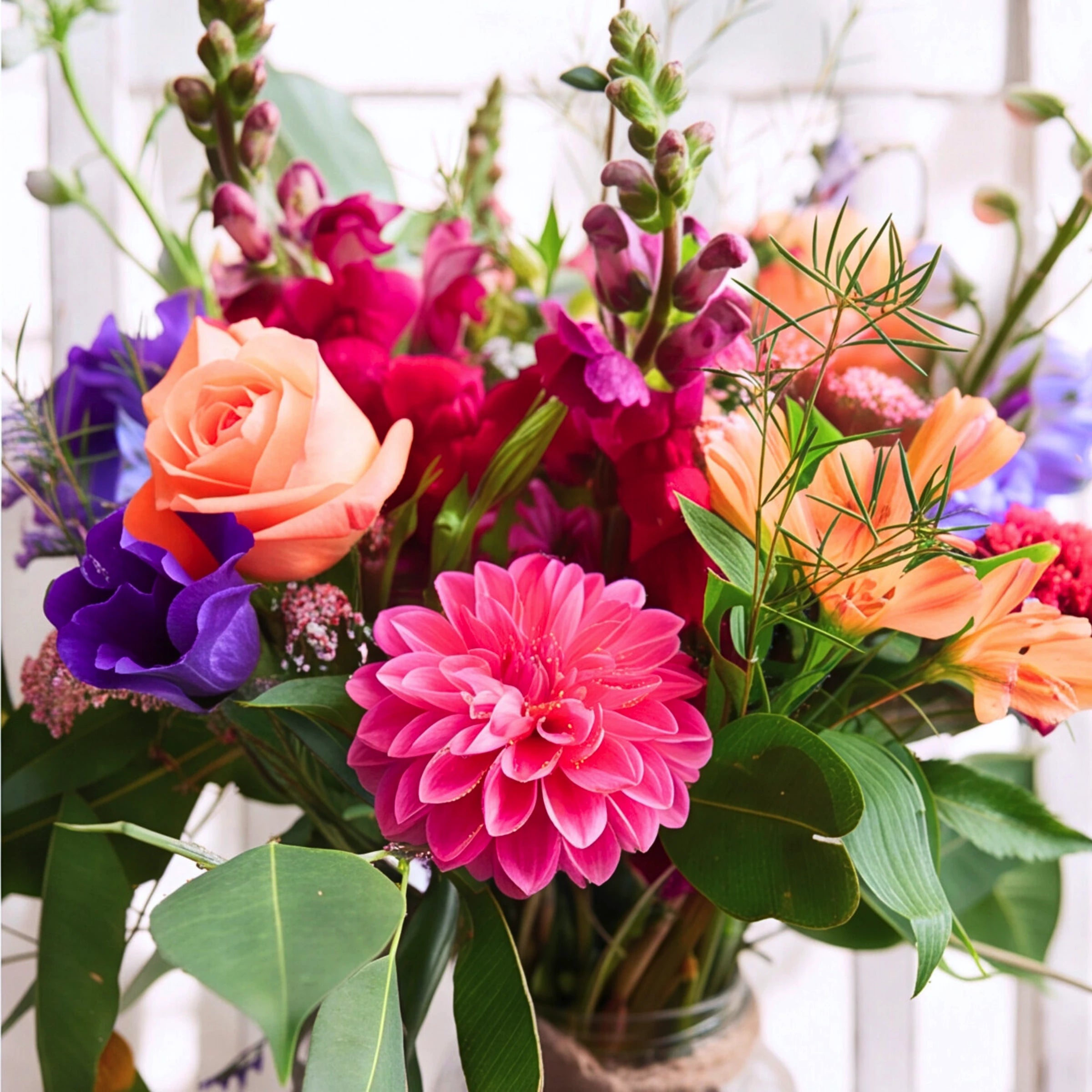 A vibrant mixed floral arrangement featuring a bright pink dahlia, peach roses, purple delphiniums, and green foliage in a glass jar vase.