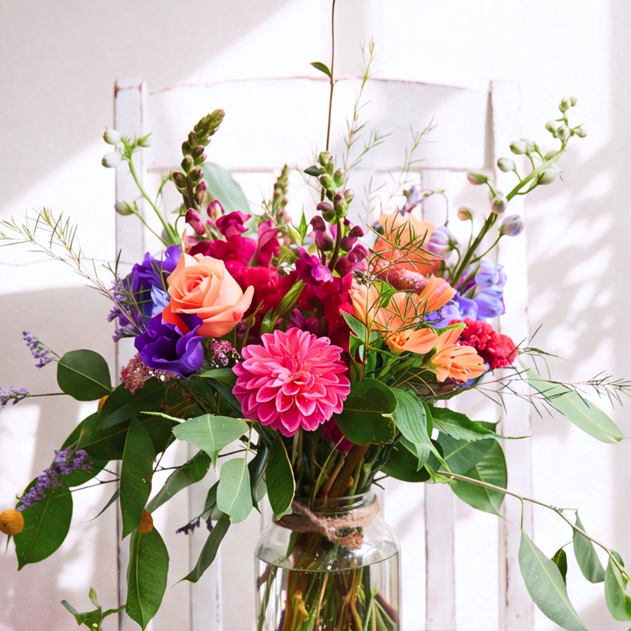 A vibrant, multicolored floral arrangement featuring pink, orange, purple, and red flowers with green foliage, presented in a clear glass jar tied with a burlap ribbon, set against a white rustic wooden chair.
