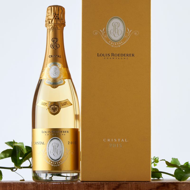 Close-up of Louis Roederer Cristal 2015 Champagne and elegant gold gift box on a wooden ledge, styled with leafy green vines against a light backdrop.