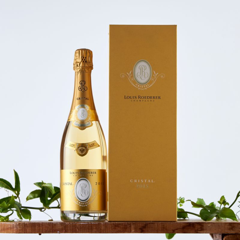 Bottle of Louis Roederer Cristal 2015 Champagne with its signature gold label and matching gift box, displayed on a wooden shelf with green foliage.
