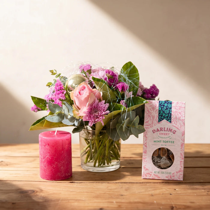 Pink rose and chrysanthemum arrangement in a small glass vase with a pink candle and a box of mint toffees on a pine table, Fabulous Flowers South Africa