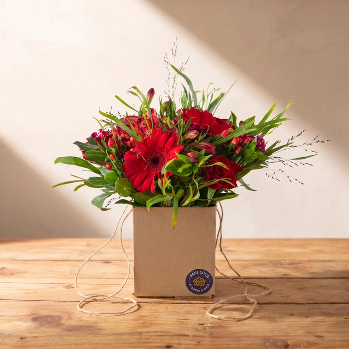 Posy of red roses, red gerberas and red alstroemeria in a small kraft box with a navy sticker on a pine table in afternoon sun, Fabulous Flowers South Africa