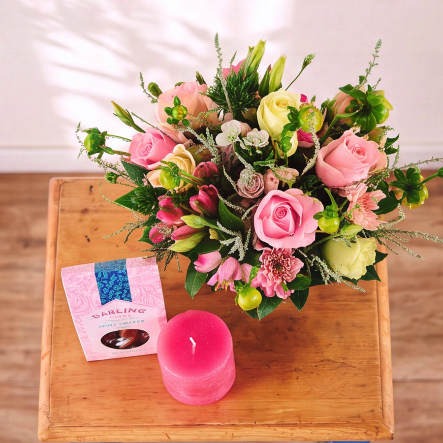 A floral bouquet with pink and pale yellow roses, green foliage, and small accent flowers arranged on a wooden tray beside a pink candle and a small pink packaged item.