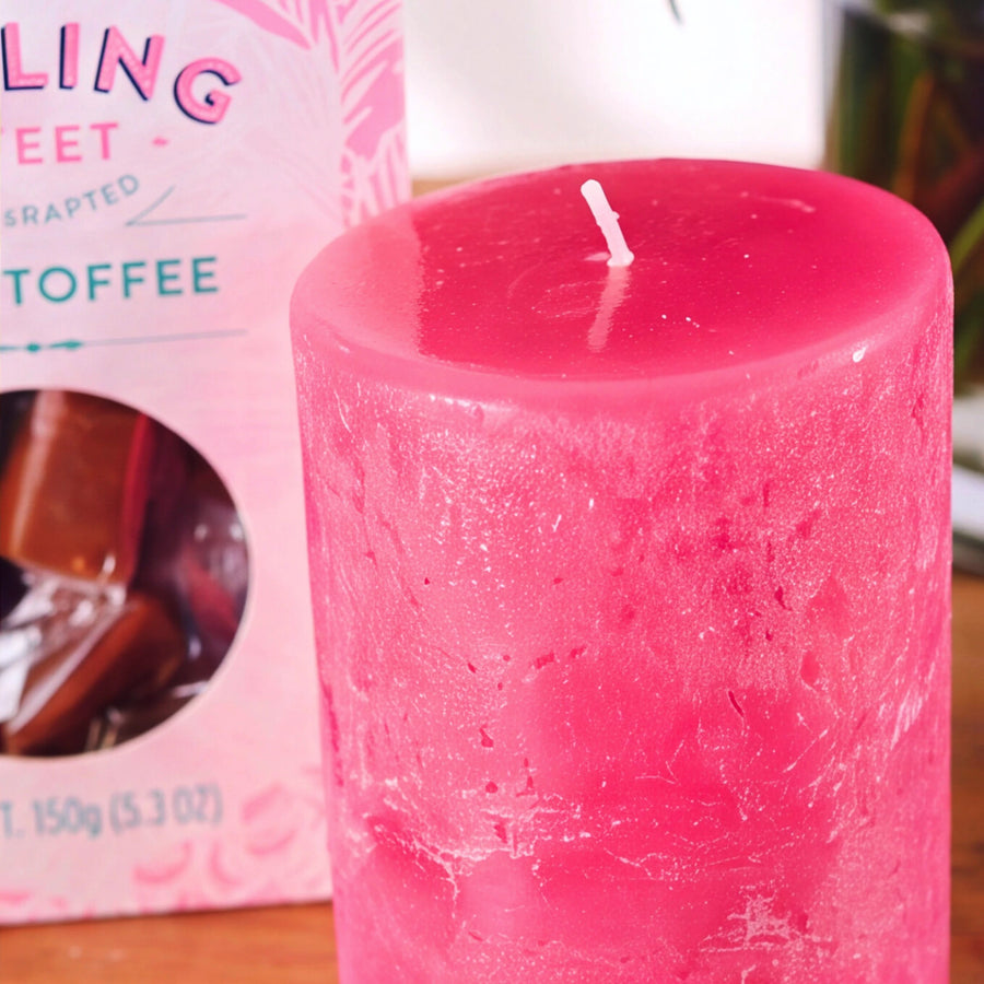 A pink textured pillar candle in the scent 'Melting Sweet Toffee' stands next to its pink packaging box on a wooden surface.
