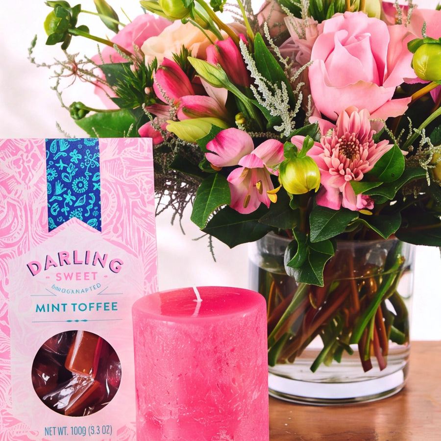 A pink candle and a pink package of Darling Sweet Mint Toffee candies are styled in front of a vibrant pink and green flower bouquet on a wooden surface.