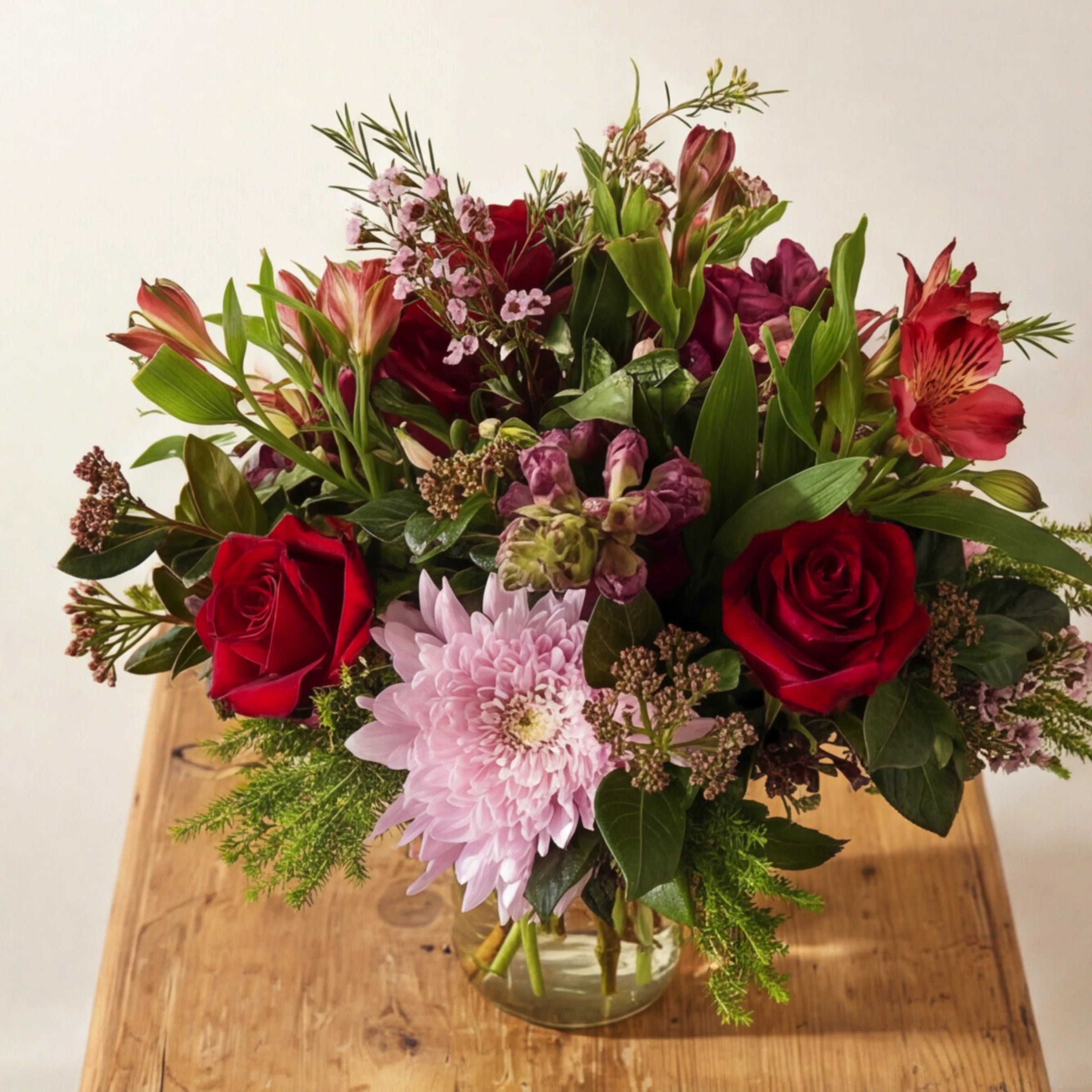 Front view of Love in a Jar romantic arrangement on a wooden stool, red roses, pink chrysanthemum, burgundy snapdragons, red and pink alstroemeria, pink wax flower and mixed fynbos-style greenery in a glass mason jar from Fabulous Flowers Cape Town