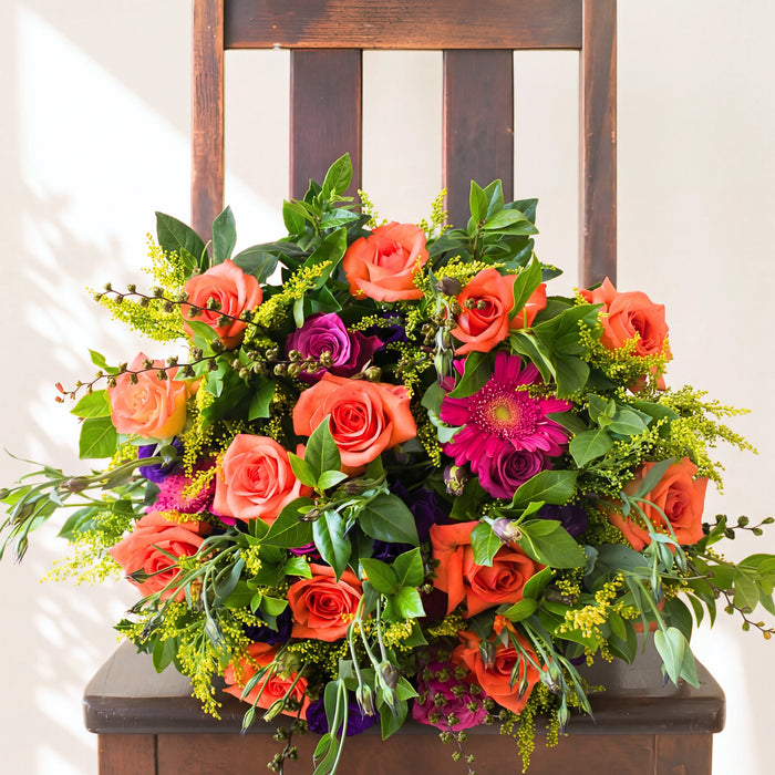 Lush and Lovely bouquet of orange roses, magenta gerberas and purple blooms with yellow solidago on a wooden chair