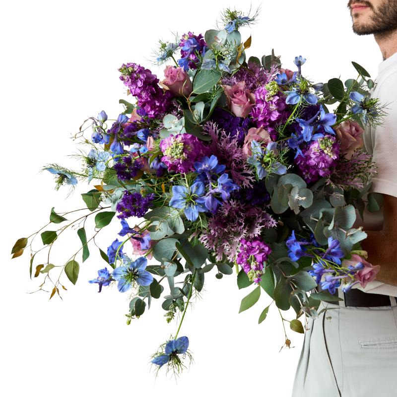 Person holding a large bouquet of purple and blue flowers with green leaves on a white background
