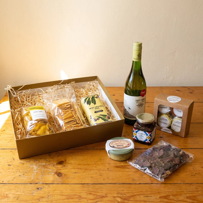 Open gold gift box of crisps, crackers and macadamia oil, with a bottle of Chenin Blanc, four cheeses, fig jam, mustard and biltong unpacked beside it on a pine table, Fabulous Flowers South Africa