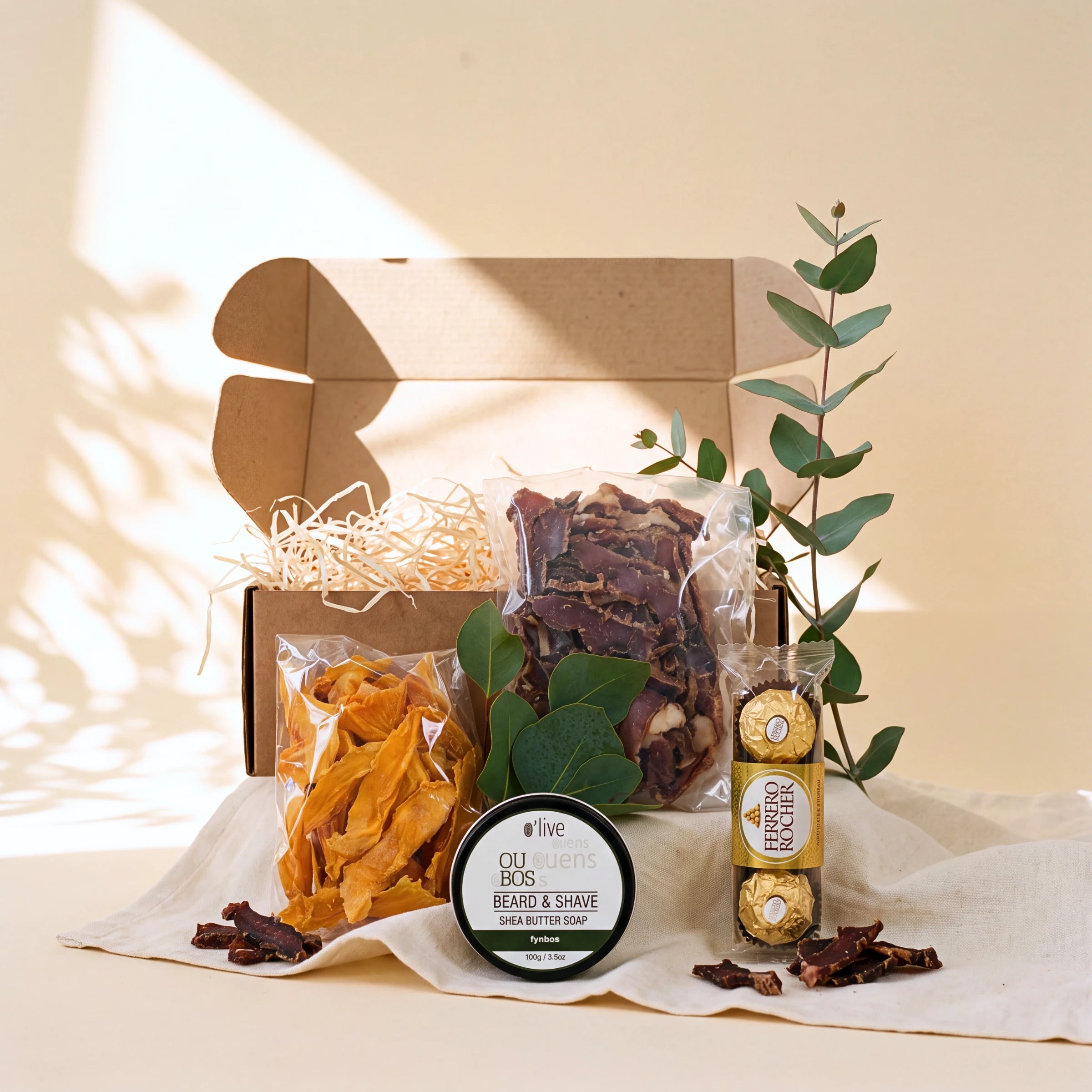A curated gift box set on a cream fabric surface with warm sunlight and leaf shadows. The set includes an open kraft cardboard box filled with shredded paper, dried orange slices in a clear bag, dark dried strips in another bag, a round black-and-white jar labeled Olive Beard & Shave, a Ferrero Rocher chocolate bar in a clear tube, and a green eucalyptus sprig.