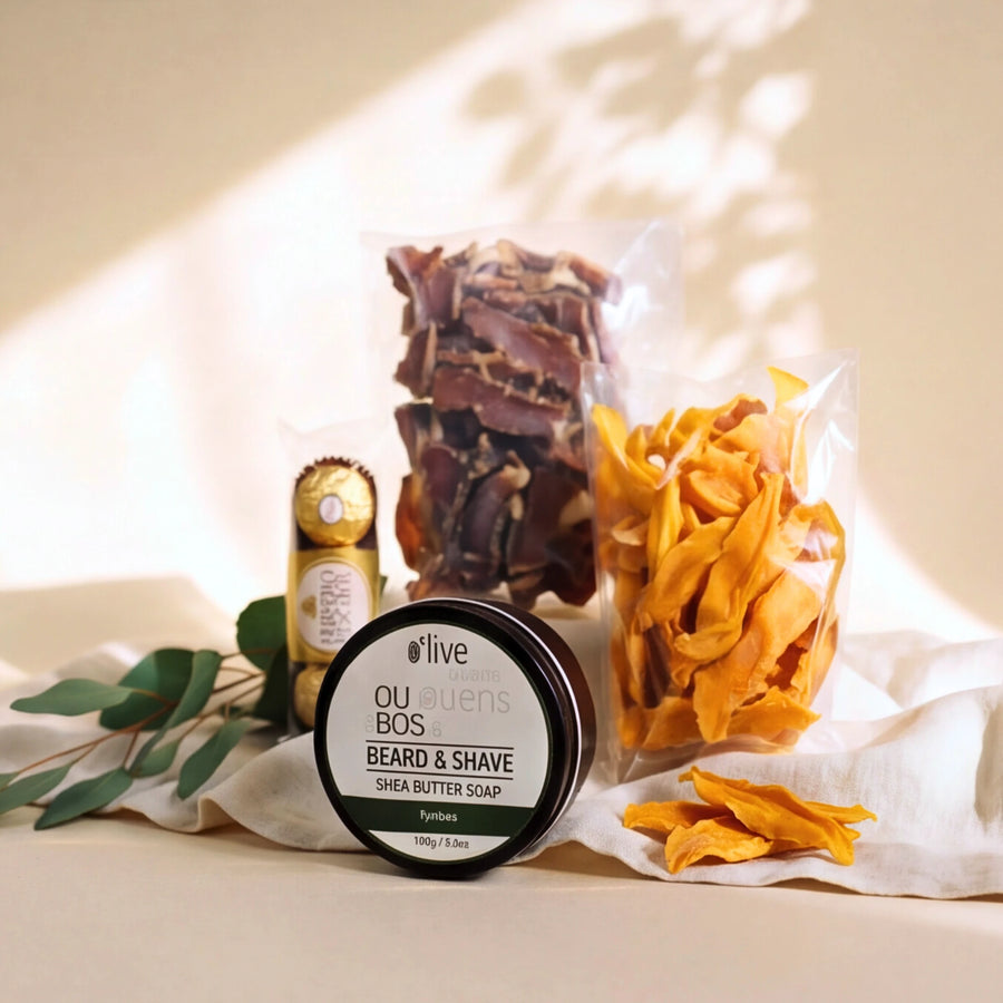 A styled product shot featuring a round tin of ou enus Beard & Shave Shea Butter Soap in the 'Tulip' scent, alongside bags of dried beard clippings and orange flower petals, with a small gold bottle, on a draped cream fabric background with soft leaf shadows.