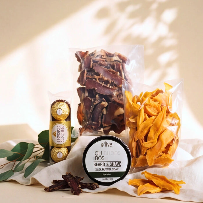 A styled product arrangement on a cream surface with soft leaf shadows. Visible items include a clear bag of dark brown dried bark pieces, a clear bag of orange dried flower or peel pieces, a round tin labeled olive Beard & Shave Bearded Shave, a wrapped Ferrero Rocher chocolate, and a small cluster of dried brown pieces in front with eucalyptus leaves nearby.