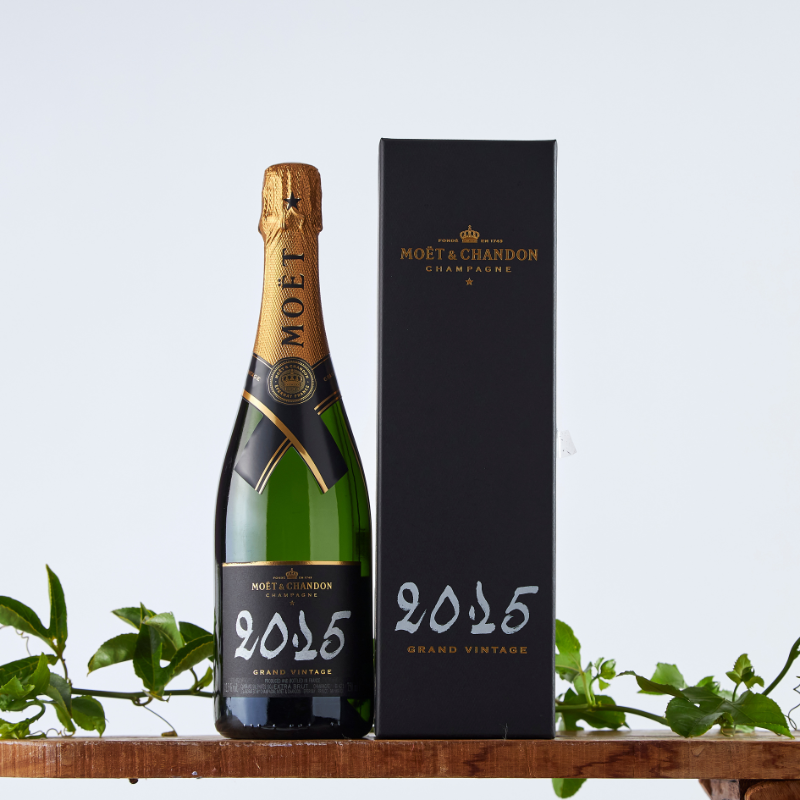 Full view of Moët & Chandon Grand Vintage 2015 Champagne and its signature black gift box with gold detailing, styled with fresh green foliage.