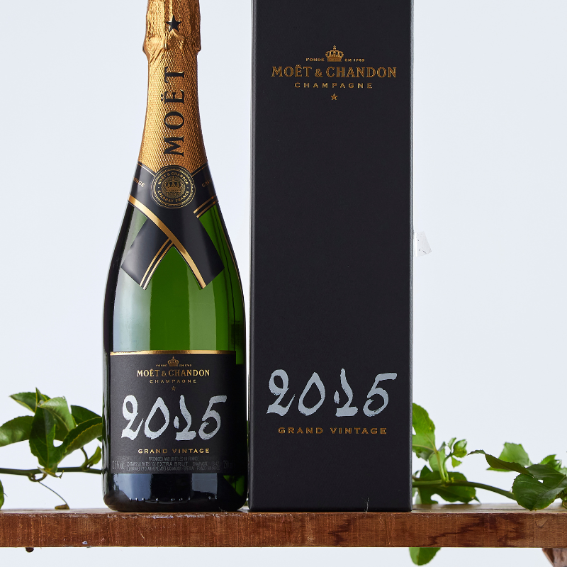 Bottle of Moët & Chandon Grand Vintage 2015 Champagne with gold foil and black label, displayed alongside its elegant black gift box on a wooden shelf with greenery.