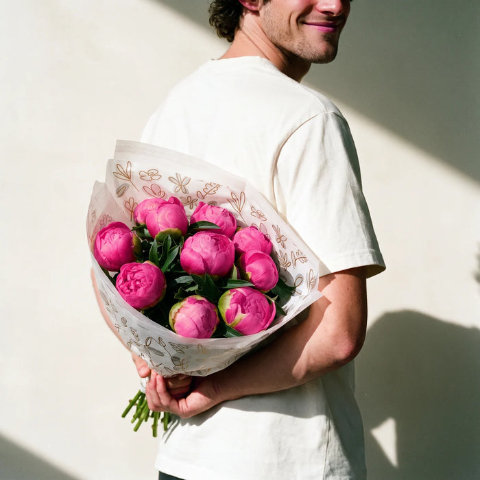 Man with curly dark hair in a white t-shirt grinning as he hides a bouquet of pink Monsieur Jules Elie peonies behind his back, Fabulous Flowers South Africa