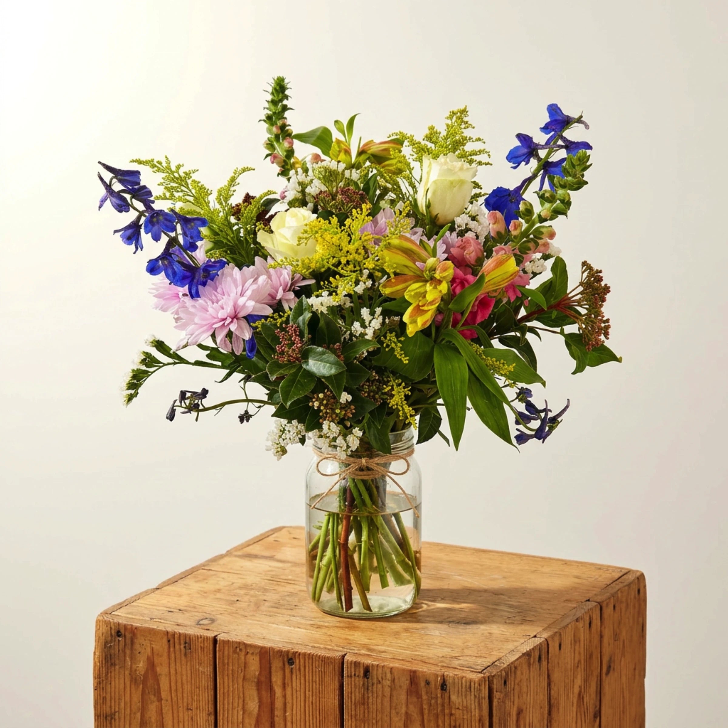 New Baby Jar arrangement on a rustic wooden block, white roses, pink chrysanthemum, blue delphinium, pink snapdragons, yellow solidago and mixed pastel greenery in a recycled glass mason jar with twine bow, Fabulous Flowers Cape Town.
