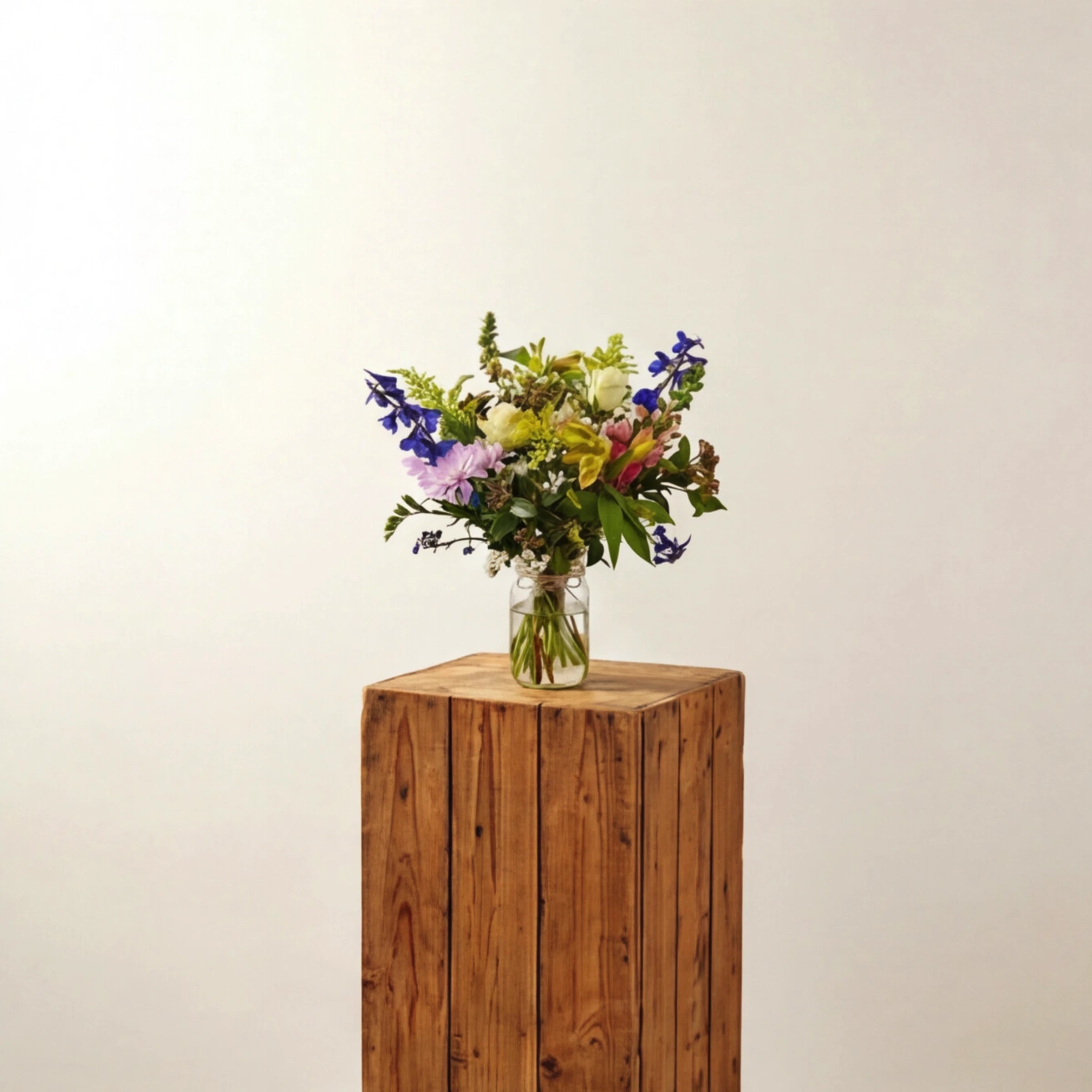 New Baby Jar arrangement on a tall wooden pedestal, small mason-jar arrangement of white roses, pink chrysanthemum, blue delphinium and pastel blooms by Fabulous Flowers Cape Town, showing the compact size for hospital and nursery delivery.