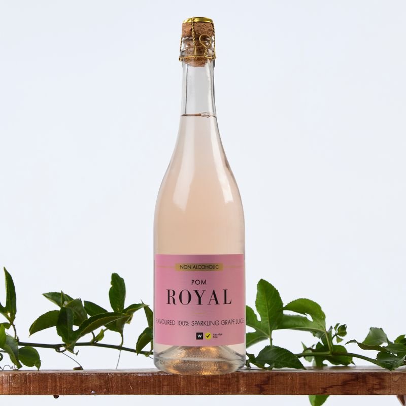 Non Alcoholic Pom Royal Sparkling 100 % Grape Juice 750 ml bottle with pink label on wooden shelf, styled with green foliage