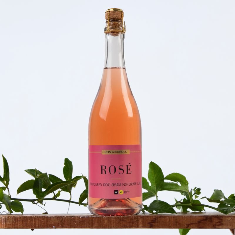 Non Alcoholic Sparkling Grape Juice 750 ml bottle with pink label on wooden shelf, styled with fresh green foliage
