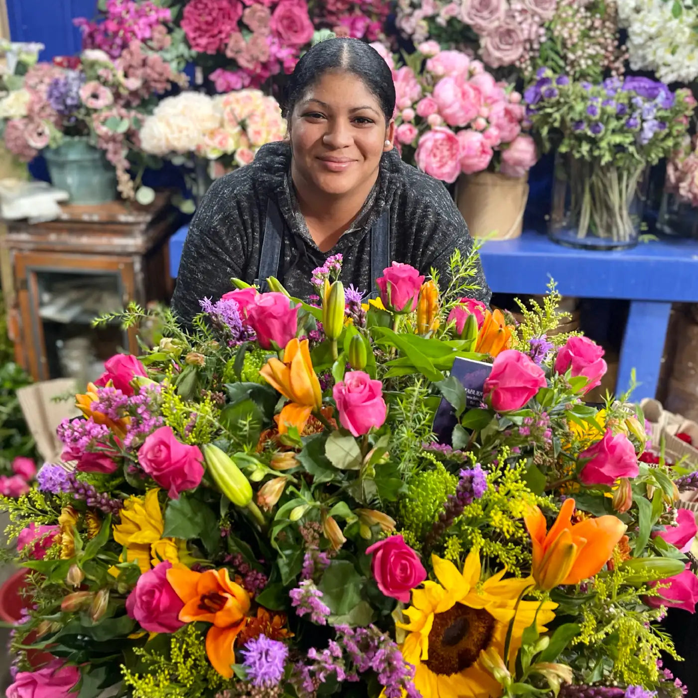 Smiling florist posing with a large, vibrant bouquet featuring pink roses, orange lilies, yellow sunflowers, and purple blooms, surrounded by other floral arrangements in a workshop. Contact us for your flower and gift needs. Fabulous Flowers and Gifts.