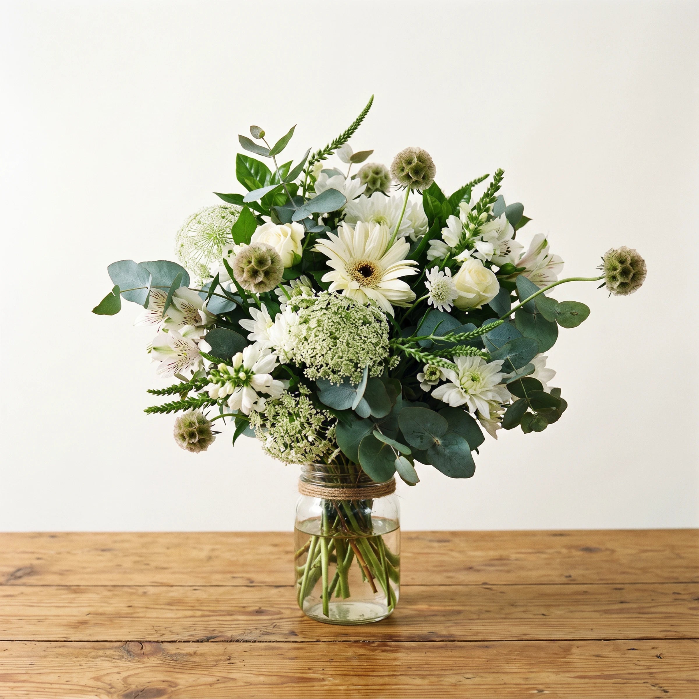 Peace in a Jar by Fabulous Flowers South Africa styled on a rustic wooden table, white sympathy flower arrangement with white roses, white gerbera, scabiosa pods, Queen Anne's lace and eucalyptus in a twine-tied glass jar