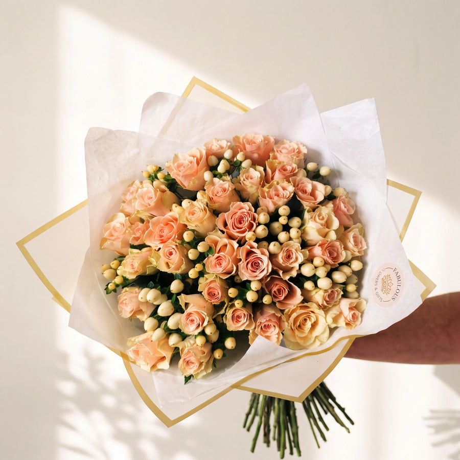 A bouquet of peach and cream roses wrapped in white tissue paper with gold borders, held by a person's arm against a warm off-white background with soft shadows.
