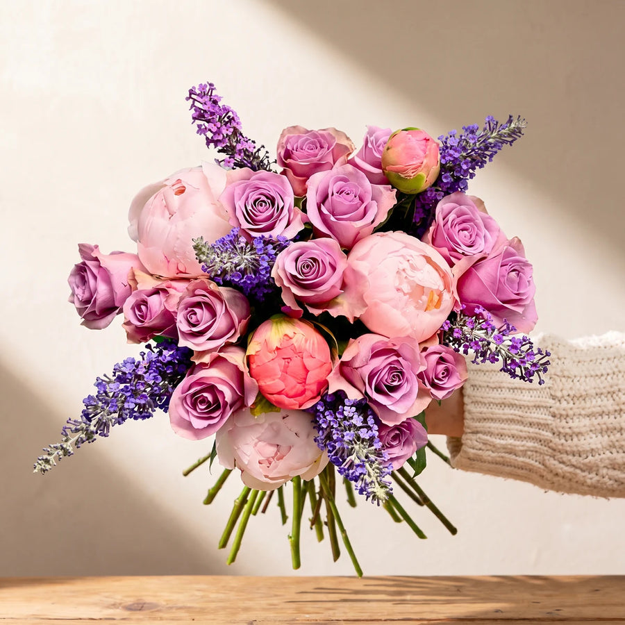 Arm holding out a bouquet of blush peonies, lilac roses and purple lavender against a sunlit wall