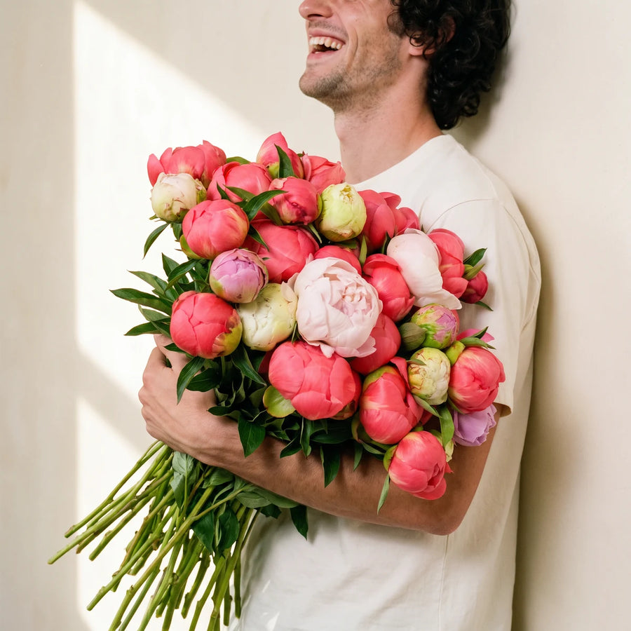 Laughing man in a white T-shirt hugging an armful of coral and pink peonies against a sunlit wall