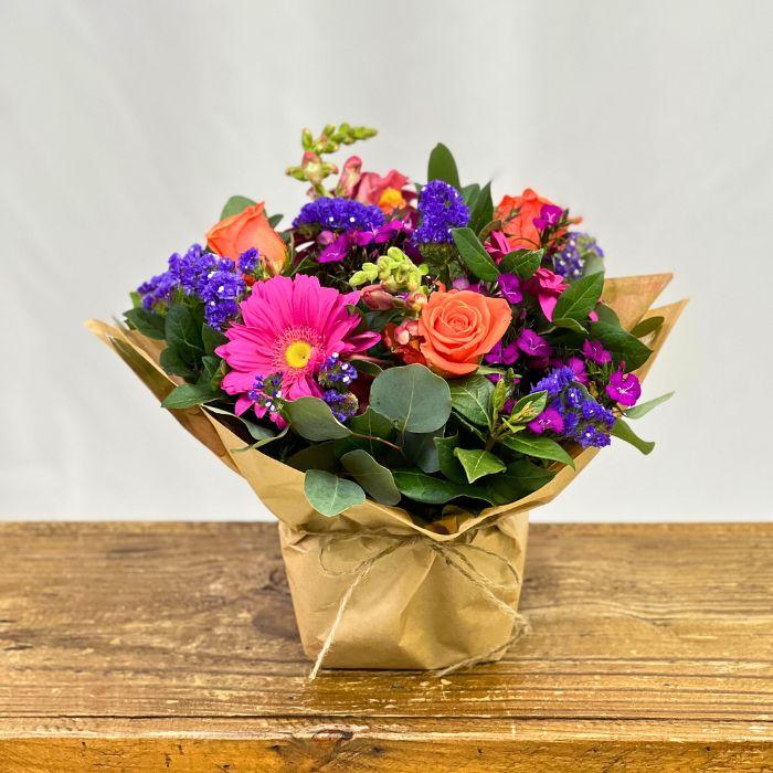Colorful bouquet of flowers in a brown paper bag on a wooden surface with a light gray background. Fabulous Flowers and Gifts.