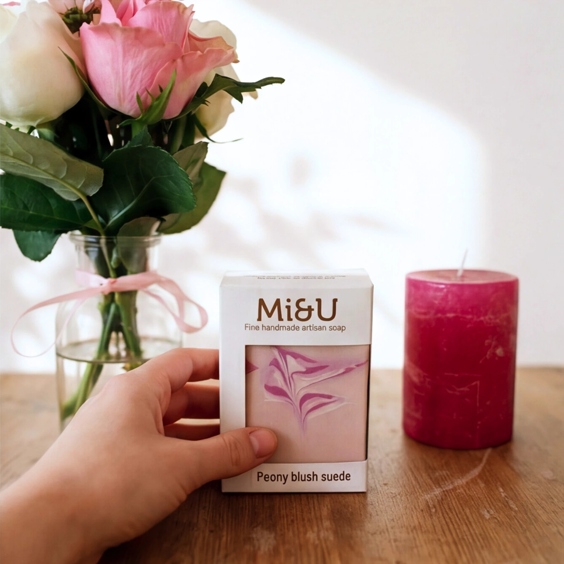 A hand holding the Mi&U Peony Blush Suede soap, pink roses and a candle behind