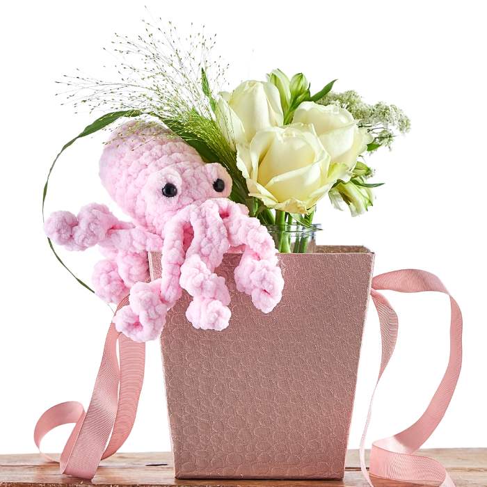 Pink knitted octopus toy in a gift bag with flowers on a white background