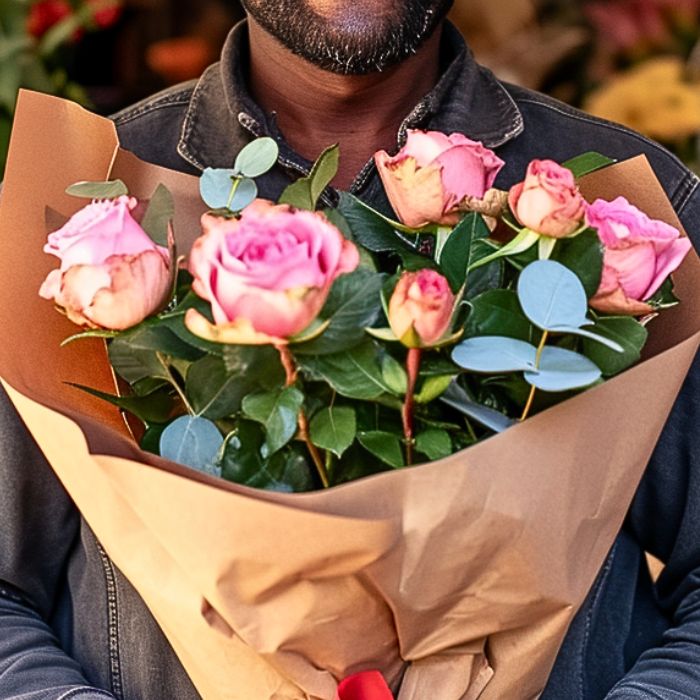 Bouquet of pink roses and green leaves wrapped in brown paper held by a person.