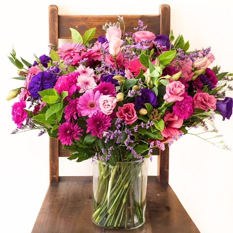 Big arrangement of colourful flowers in a clear vase on a wooden chair.