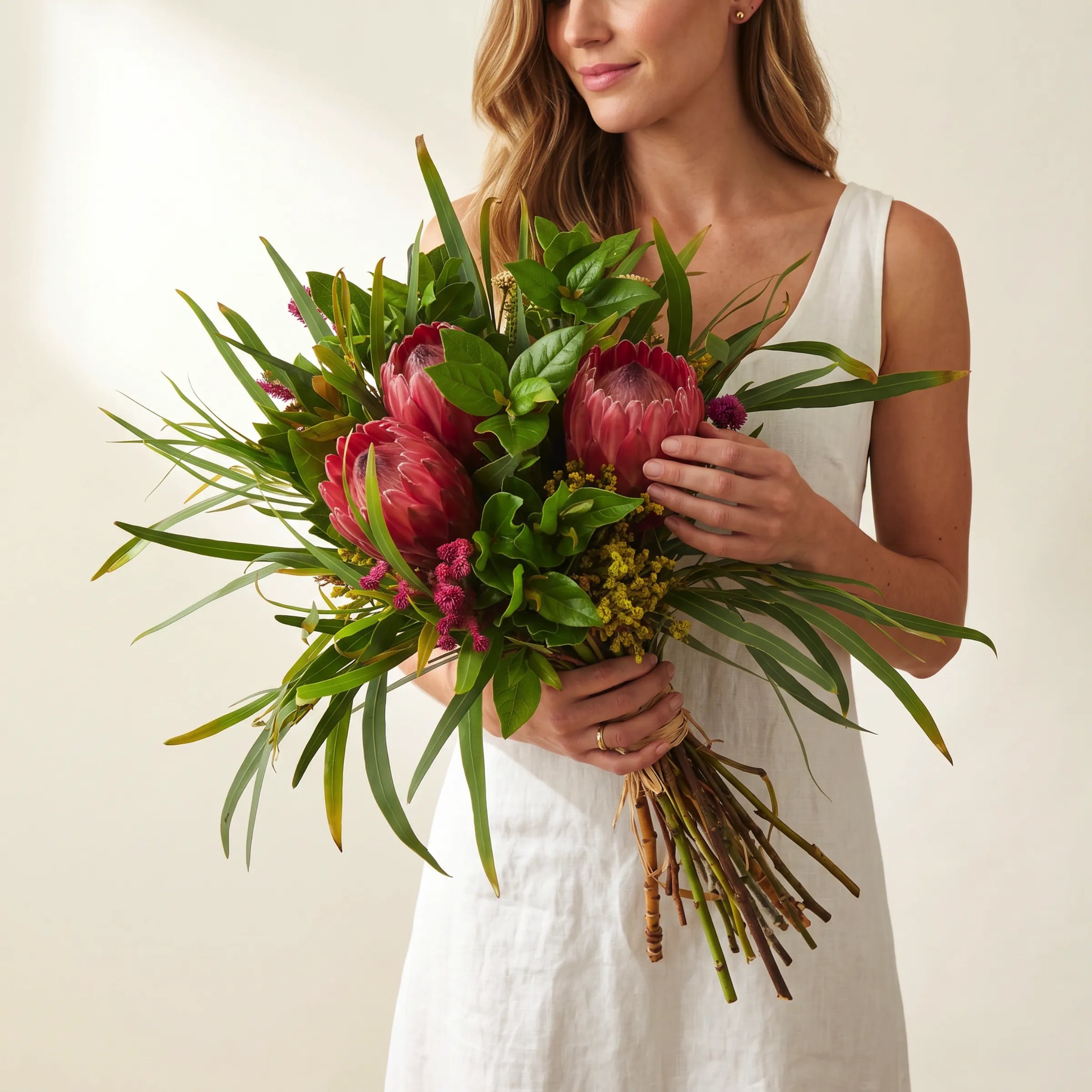 Protea Garden Flower Bouquet held by a woman in a white linen dress, hand-tied arrangement of three pink king proteas, long strappy eucalyptus leaves, magenta celosia, yellow solidago and lush green pittosporum foliage tied with raffia, Fabulous Flowers South Africa