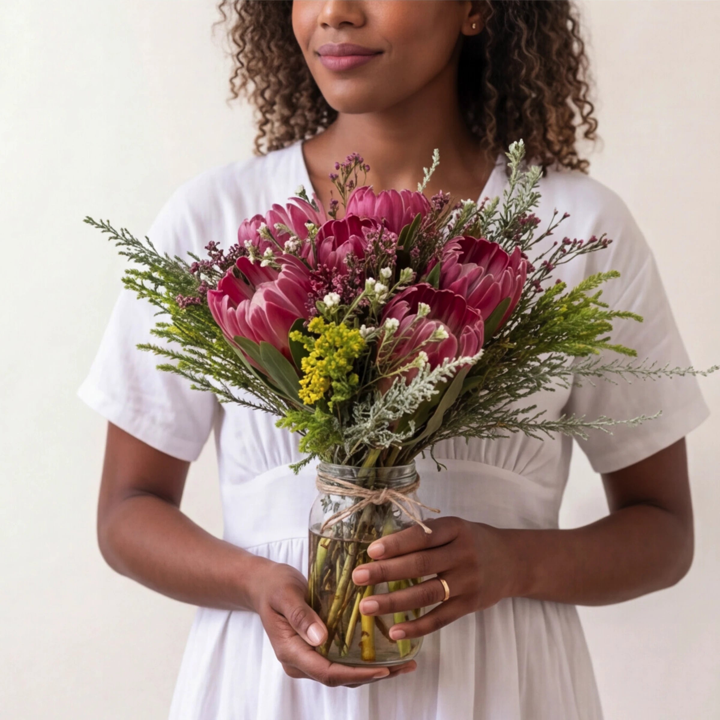 Woman in white holding Proteas in a Jar from Fabulous Flowers South Africa, Pink Ice proteas, pink fynbos waxflower, solidago, restio and silver wormwood in a twine-tied glass jar for same-day Cape Town delivery