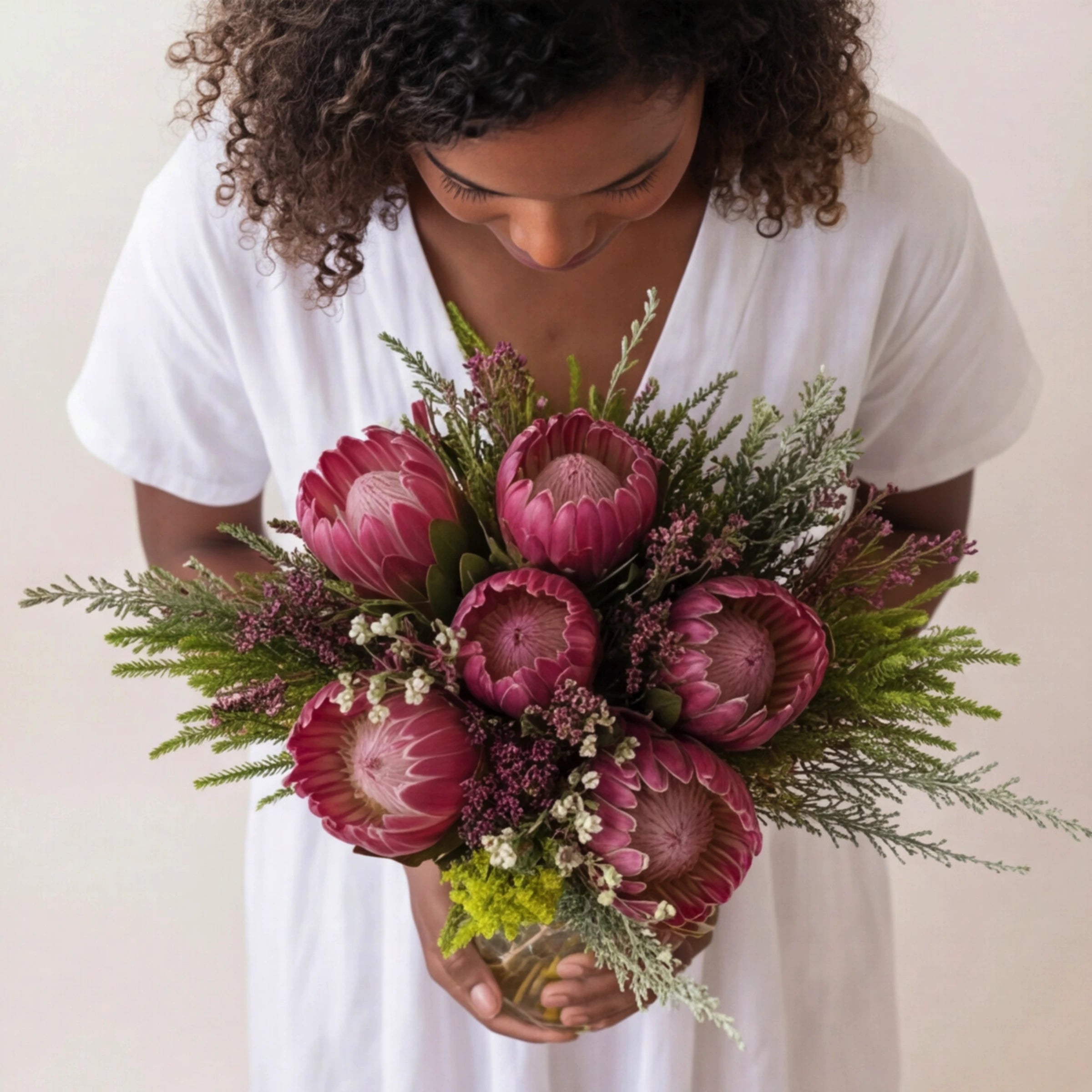 Woman holding Proteas in a Jar by Fabulous Flowers South Africa close to her chest, Pink Ice proteas, fynbos waxflower, erica and restio hand-styled in a twine-tied glass jar, a South African indigenous flower arrangement