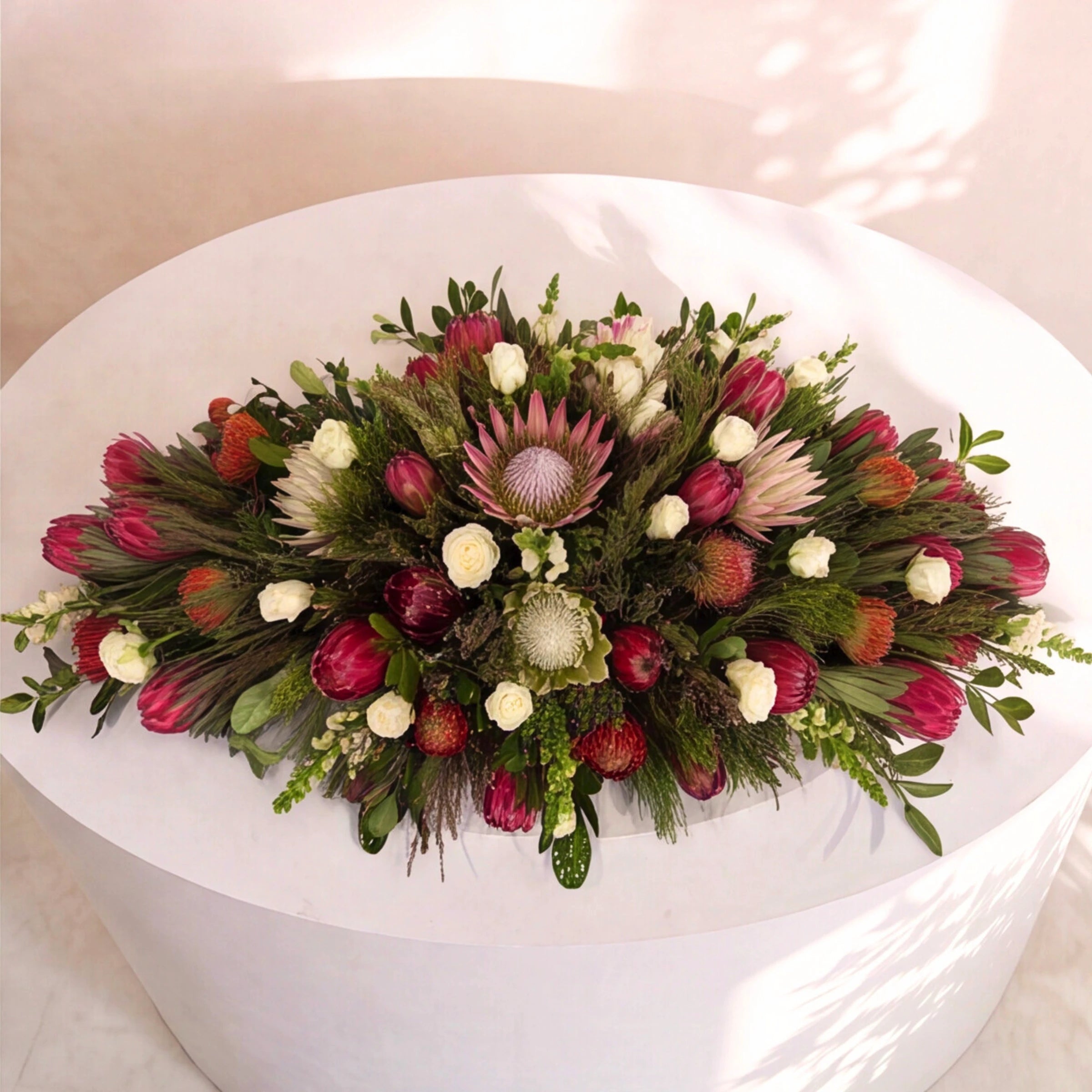 A large, vibrant floral arrangement featuring pink protea-like flowers, deep red roses, white daisies, and green foliage, displayed on a round white pedestal.