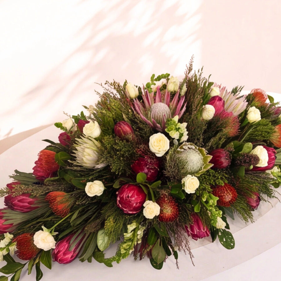A lush, long floral arrangement featuring a mix of red protea, white roses, green foliage, and pink protea blossoms on a light-colored surface.