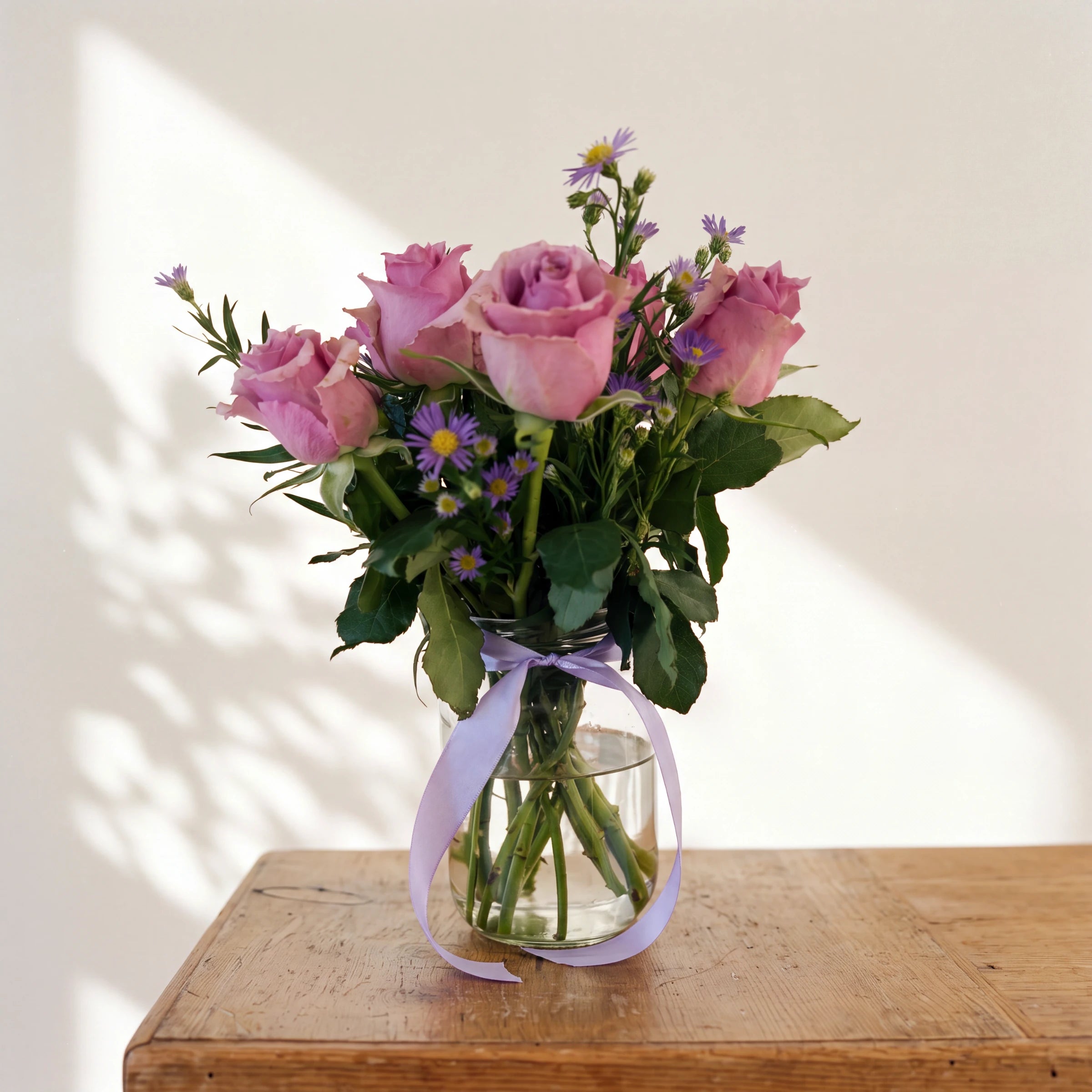 Provence lavender rose arrangement of mauve roses and purple asters in a glass vase with a lilac ribbon on a wooden table